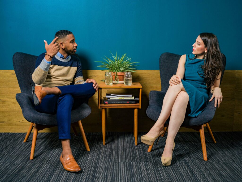 A man and woman engaged in conversation, sitting in stylish armchairs against a blue wall with a wooden side table and potted plants.