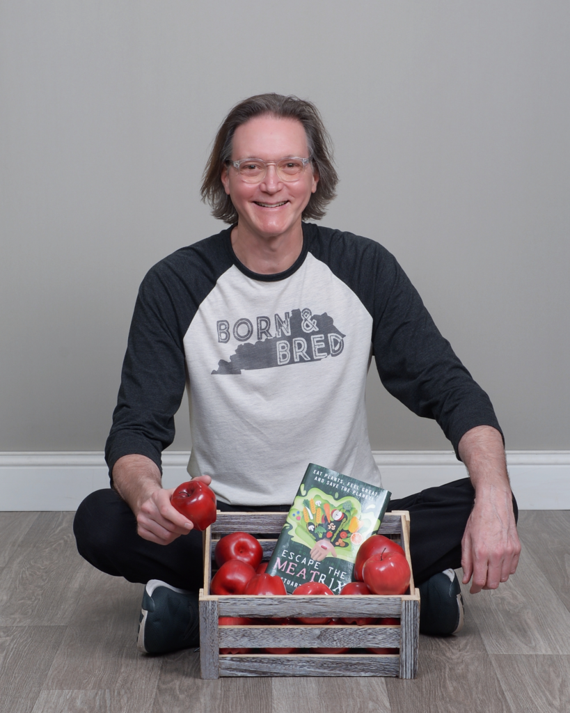 Author Stuart Waldner seated on the floor smiling, holding a red apple in one hand with his book 'Escape the Meatrix' in a crate of apples, highlighting plant-based nutrition.
